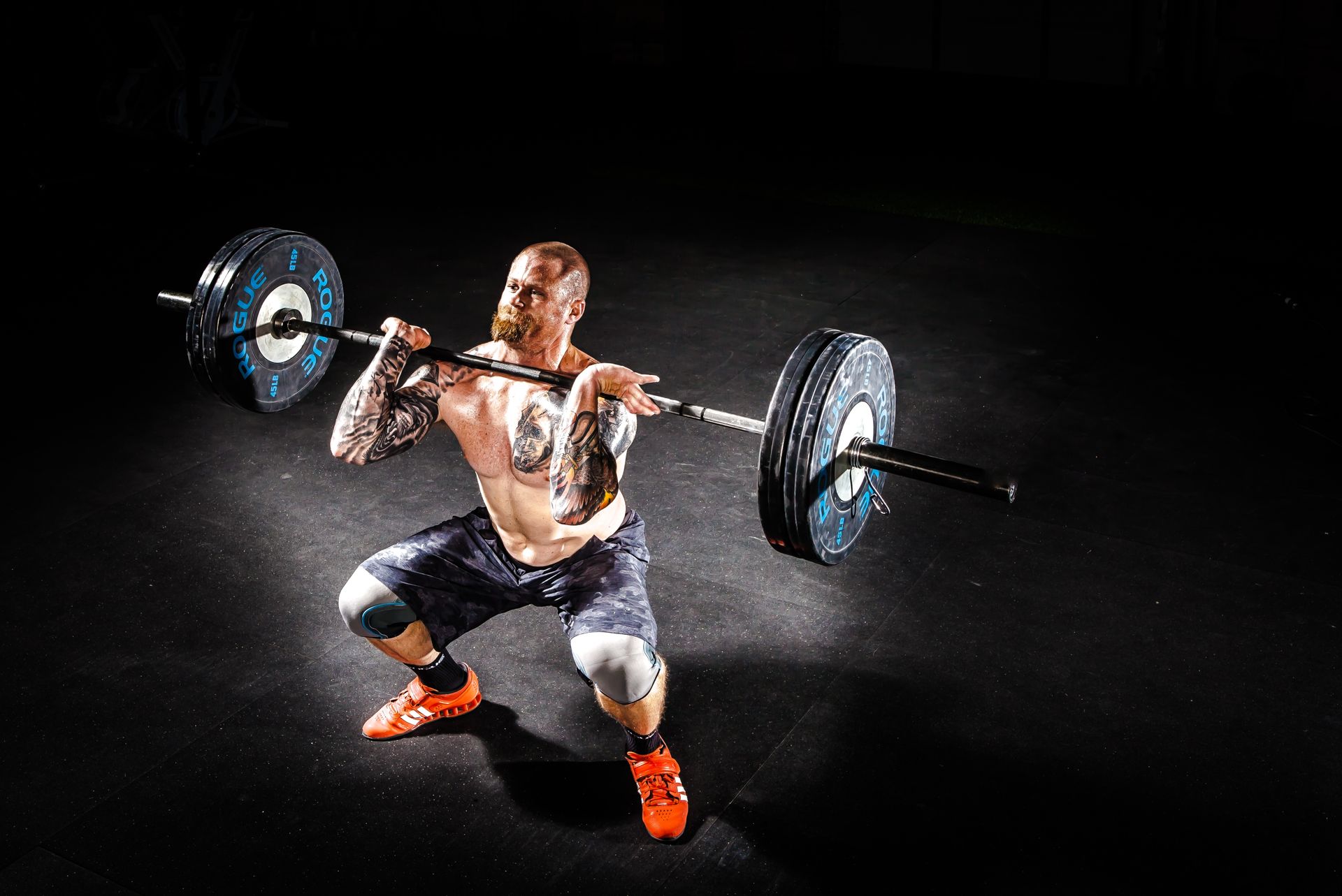 Lifter mid front squat in a dark garage gym, barbell resting across the front delts with elbows high, chalk dust in the air